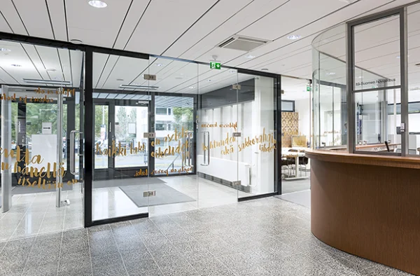 Kuopion oikeustalon vastaanottotiski ja kaareva lasiseivä sanataideteuksella. Suunnitellut Partanen & Lamusuo Oy | The reception desk and curved glass wall with a literary art piece at the Kuopio Courthouse, designed by Partanen & Lamusuo Ltd.